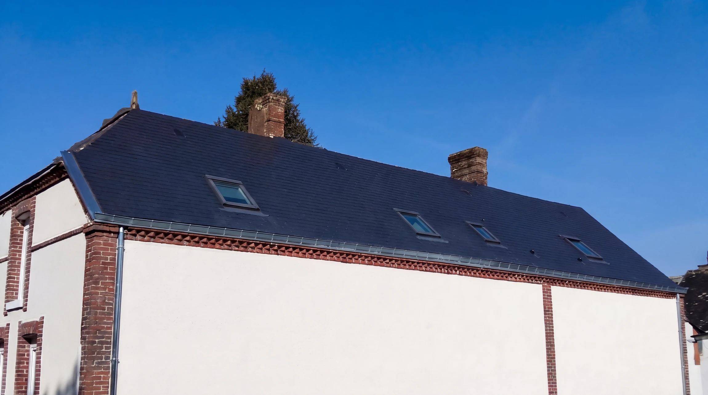 Toiture en ardoise naturelle noire, vue en contre-plongée, sur bâtisse ancienne à murs enduits blancs et encadrements en briques rouges apparentes